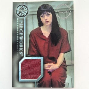 May include: A trading card featuring a woman in a red outfit. The card has the text "PIECEWORKS" and "inkworks" printed on it. A textured red fabric swatch is embedded in the card.