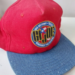Könnte beinhalten: Rote und blaue Baseballkappe mit einem gestickten Aufnäher in Gelb, Blau und Schwarz, auf dem "San Antonio, Texas, G.J.J.E. Remember the Legend" steht. Der Aufnäher zeigt einen weißen Stern in einem roten Umriss.