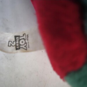 May include: A close-up of a plush toy with a red and green color scheme. The toy has a tag with the text "Toy Story 2" printed on it. The background is a light, off-white color.