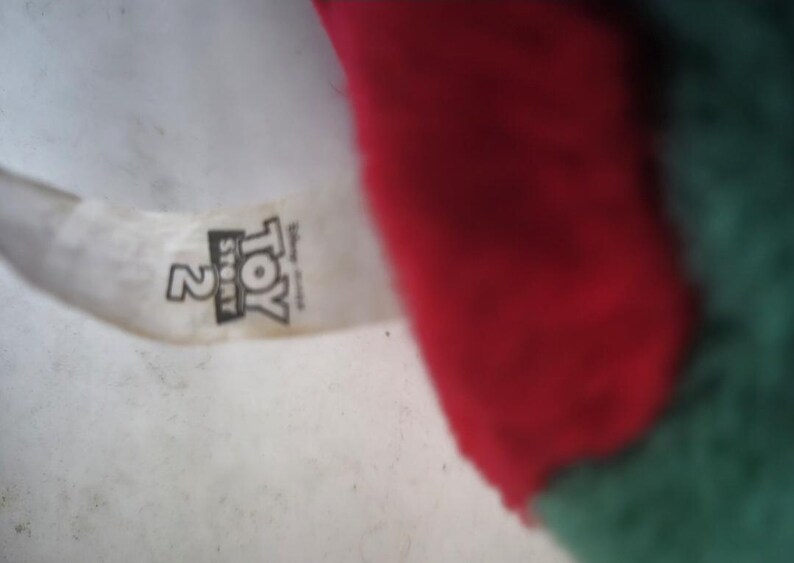 May include: A close-up of a plush toy with a red and green color scheme. The toy has a tag with the text "Toy Story 2" printed on it. The background is a light, off-white color.