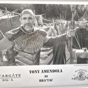 May include: Monochrome photograph of Tony Amendola as Bra'tac from Stargate SG-1. Amendola is in costume, featuring a headpiece, armour, and a cape, holding a staff. The image includes the Stargate SG-1 logo and the Metro-Goldwyn-Mayer trademark.