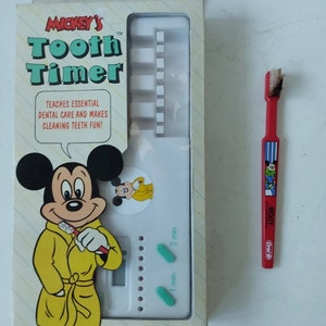 Può includere: Un timer per denti Mickey Mouse bianco con un'immagine di Mickey Mouse che si spazzola i denti. Il timer ha un quadrante con impostazioni di 1 e 2 minuti. La scatola include anche uno spazzolino da denti rosso con un design di Mickey Mouse.