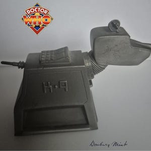 Op de afbeelding: Een metallic grijze H-9 robot hond figuur uit de Doctor Who serie. Het beeldje heeft een hoekig lichaam, een kop met een rond oor en een lange spuitmond. Het Doctor Who logo is zichtbaar in de linkerbovenhoek.