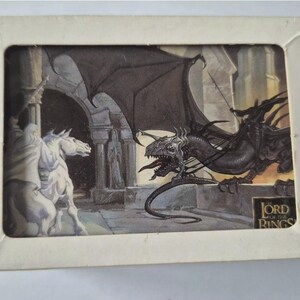 Puede incluir: Una caja rectangular de color blanco roto con una ventana que muestra una ilustración de fantasía. La imagen representa a una figura sobre un caballo blanco frente a una criatura alada oscura con un jinete. El texto The Lord of the Rings aparece abajo.
