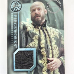 May include: A trading card featuring a man with a bald head and a beard, wearing a dark green and gold patterned jacket. The card includes the text "PIECEWORKS" and a small fabric swatch.