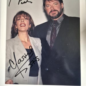 May include: A photograph featuring two people, a man and a woman, posing together. The woman wears a light-colored suit, while the man is in a dark suit with a patterned tie. The image is signed by both individuals. The text "JONATHAN & MARINA" is visible at the bottom.