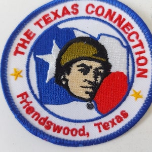 Könnte beinhalten: Ein blauer und weißer gestickter Aufnäher mit dem Text "The Texas Connection Friendswood, Texas" und einem Cartoon-Bild einer Person mit einem grünen Hut, die ein rotes Herz hält.