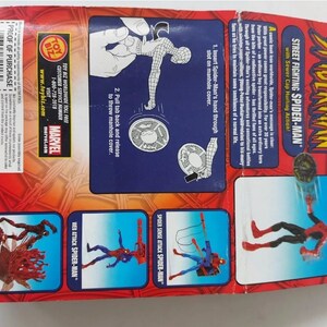 Pode incluir: Uma figura de a&ccedil;&atilde;o vermelha e preta do Homem-Aranha com uma tampa de esgoto na m&atilde;o. A figura &eacute; embalada em uma caixa de papel&atilde;o vermelha e azul com as palavras "Street Fighting Spider-Man" e "Sewer Cap Hurling Action!" impressas nela. A parte de tr&aacute;s da caixa mostra diferentes poses de a&ccedil;&atilde;o da figura.