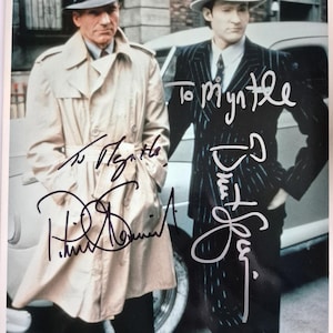 May include: A vintage photograph featuring two men in suits and fedora hats, standing next to a classic car. The image is signed with handwritten inscriptions. One man wears a light-colored trench coat.