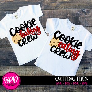 May include: Two white t-shirts with the phrases "Cookie Baking Crew" and "Cookie Eating Crew" in black and red lettering. Each shirt features a chocolate chip cookie graphic. The shirts are on a wooden surface.