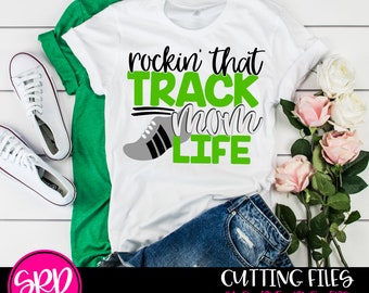 May include: White t-shirt with green text that reads "rockin' that TRACK mom LIFE" and a black and gray running shoe graphic. The shirt is laying on a green surface with a pair of white sneakers and a pair of blue jeans.