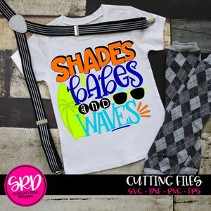 May include: A white t-shirt with the text "Shades Babes and Waves" in colorful letters. The text is surrounded by a surfboard, palm tree, and sunglasses. The shirt is paired with black suspenders and gray and white checkered pants.
