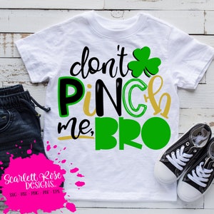 May include: White t-shirt with the text "don't pinch me, BRO" in black, green, and gold. A green shamrock is above the word "pinch". Black and white sneakers and blue pants are next to the shirt.