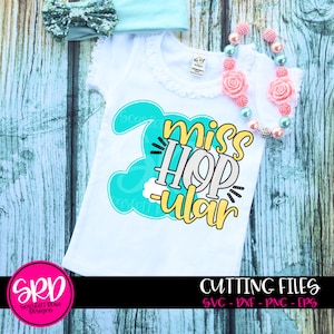 May include: A white shirt with a turquoise bunny design and the text "Miss Hop-ular" in yellow and black lettering. The shirt is on a wooden background with a pink and turquoise necklace.