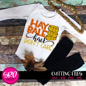 May include: White baby onesie with the text "HAY BALE hair DON'T CARE" in orange and black lettering. A stack of hay bales is pictured next to the text. The onesie is on a wooden surface with fall leaves and a pine cone.
