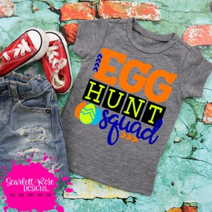 May include: A gray t-shirt with the words "EGG HUNT squad" in orange, black, and blue. The shirt is next to a pair of red sneakers and denim shorts. The shirt has a graphic of two Easter eggs.