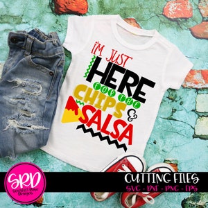 May include: A white t-shirt with a black, green, and red graphic that says "I'm Just Here For The Chips & Salsa". The shirt is on a blue and green background with a pair of jeans and red shoes.