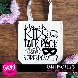 Op de afbeelding: Een witte tote bag met zwarte tekst die "Ik leer kinderen terugpraten wat is jouw superpower?" zegt met een zwarte superheldenmasker afbeelding. De tas ligt op een houten oppervlak met roze bloemen.