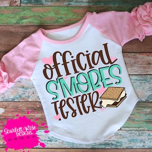 May include: A white and pink long-sleeved shirt with the text "official s'mores tester" in brown and teal lettering. There are small pink hearts and a cartoon image of a s'more on the shirt.