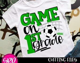 May include: White t-shirt with the text "GAME ON 1ST GRADE" in green and black, featuring a soccer ball graphic. The shirt also has a small green arrow design. The shirt is a children's apparel item.