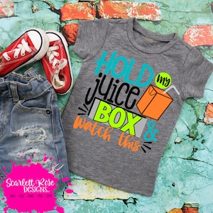 May include: A gray t-shirt with the text "Hold my juice box & watch this" in colorful letters. The text is surrounded by a stylized juice box with a straw.
