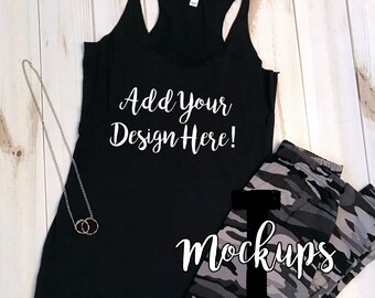 Puede incluir: Camiseta de tirantes negra con el texto "Add Your Design Here!" impreso en blanco. La camiseta está sobre una superficie de madera blanca con un collar de cadena plateada y unos leggings de camuflaje.