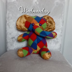 May include: A colorful patchwork elephant plush toy with gold satin ears. The elephant is sitting on a white fluffy surface. The text "Wednesday" is written above the elephant.