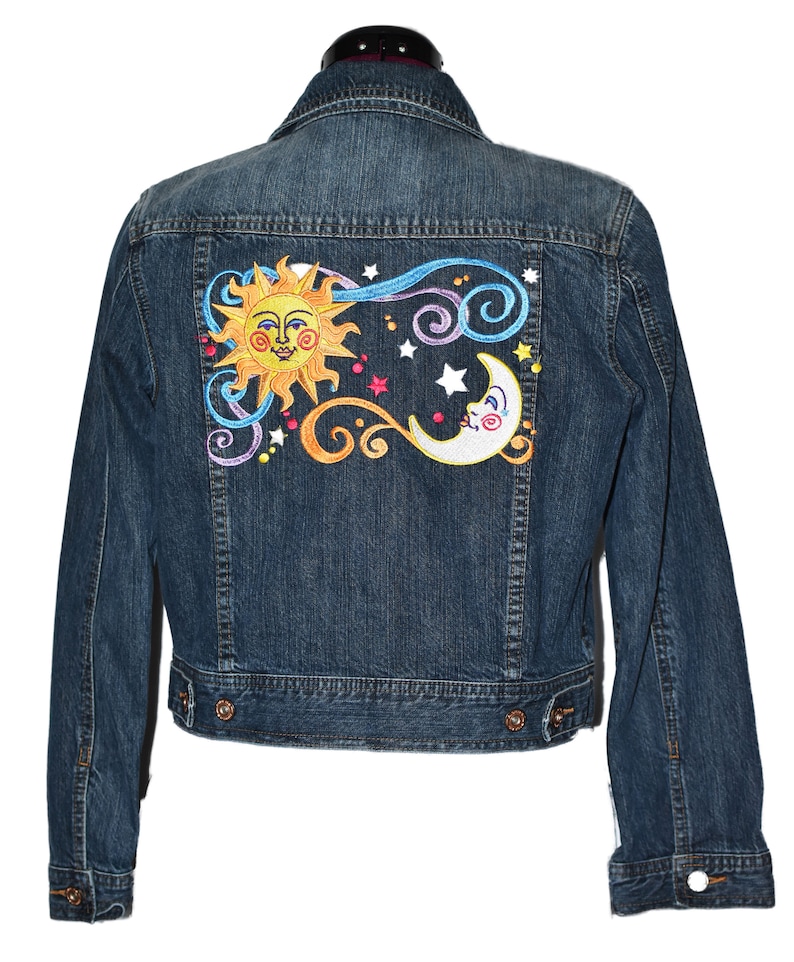 Puede incluir: Una chaqueta vaquera azul con un dise&ntilde;o de sol y luna bordado en la espalda. El sol es amarillo con una cara sonriente y la luna es blanca con forma de media luna.