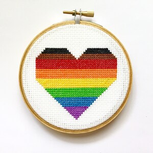 Peut inclure: Un cœur en point de croix dans un cercle à broder en bois. Le cœur est composé de rayures horizontales aux couleurs du drapeau de la fierté, dont le marron, le noir, le rouge, l'orange, le jaune, le vert, le bleu et le violet. Le fond est blanc.