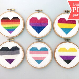 May include: Six embroidery hoops with white fabric and different coloured heart designs. The hearts are in the style of various pride flags, including rainbow, bisexual, pansexual, genderqueer, transgender, and non-binary.