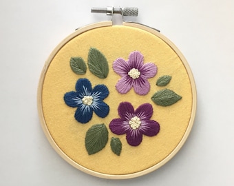 May include: Embroidered floral hoop art featuring three flowers in shades of blue, purple, and violet, with green leaves, on a yellow fabric. The embroidery is set in a wooden hoop.
