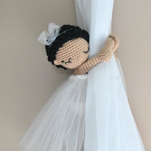 May include: A crocheted ballerina doll with a white tulle skirt and a white bow in her hair is holding a white curtain. The doll has brown eyes and black hair. The doll is a curtain tieback.