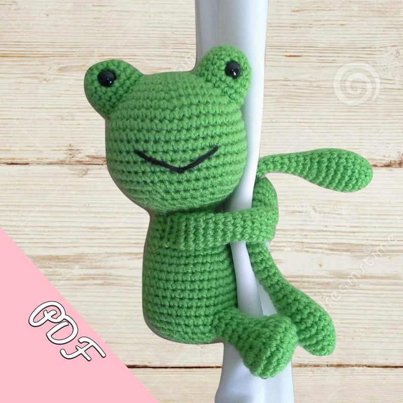 May include: Green crocheted frog toy with black eyes and a smile, holding onto a white pole. The frog is hanging from the pole with its arms and legs wrapped around it. The background is a light wood surface.