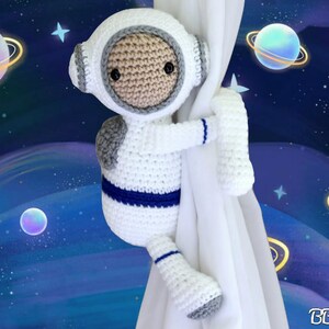May include: A white crocheted astronaut toy with blue accents holding onto a white curtain. The background is a blue and purple space scene with planets and stars.