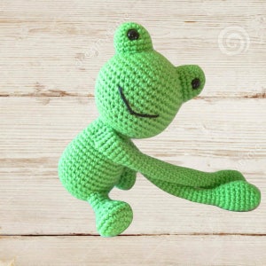 May include: A green crocheted frog toy with black eyes and a smiling mouth. The frog is sitting on a light wood background.