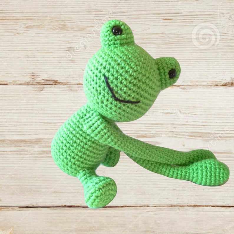 May include: A green crocheted frog toy with black eyes and a smiling mouth. The frog is sitting on a light wood background.