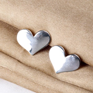 May include: A pair of silver heart-shaped earrings. The earrings are smooth and shiny, and they are lying on a brown fabric surface.