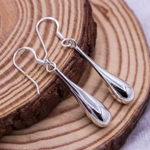 Può includere: Un paio di orecchini a goccia in argento lucido. Gli orecchini hanno una superficie liscia e riflettente e un design semplice ed elegante. Sono appesi a un gancio curvo. Gli orecchini sono esposti su una superficie di legno.