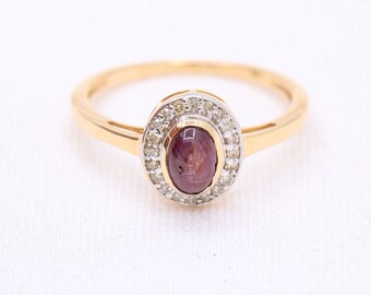 May include: A gold ring featuring a red oval gemstone set within a halo of smaller white gemstones.