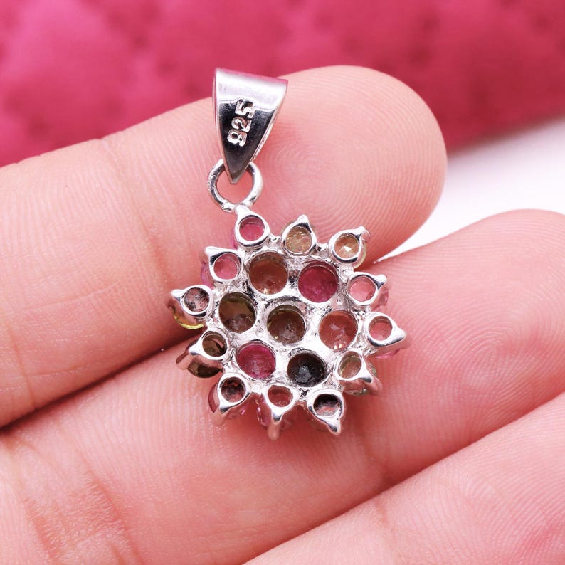 May include: A silver pendant with a floral design, featuring a cluster of colorful gemstones. The pendant has a loop at the top for a chain and is held in a hand. The gemstones are various shades of pink, brown, and green.