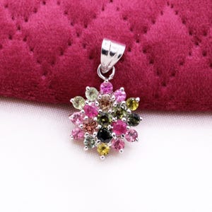 May include: A silver pendant with a cluster of colorful gemstones. The gemstones are arranged in a flower-like pattern, featuring shades of pink, green, yellow, and black. The pendant is set against a deep red, quilted background.