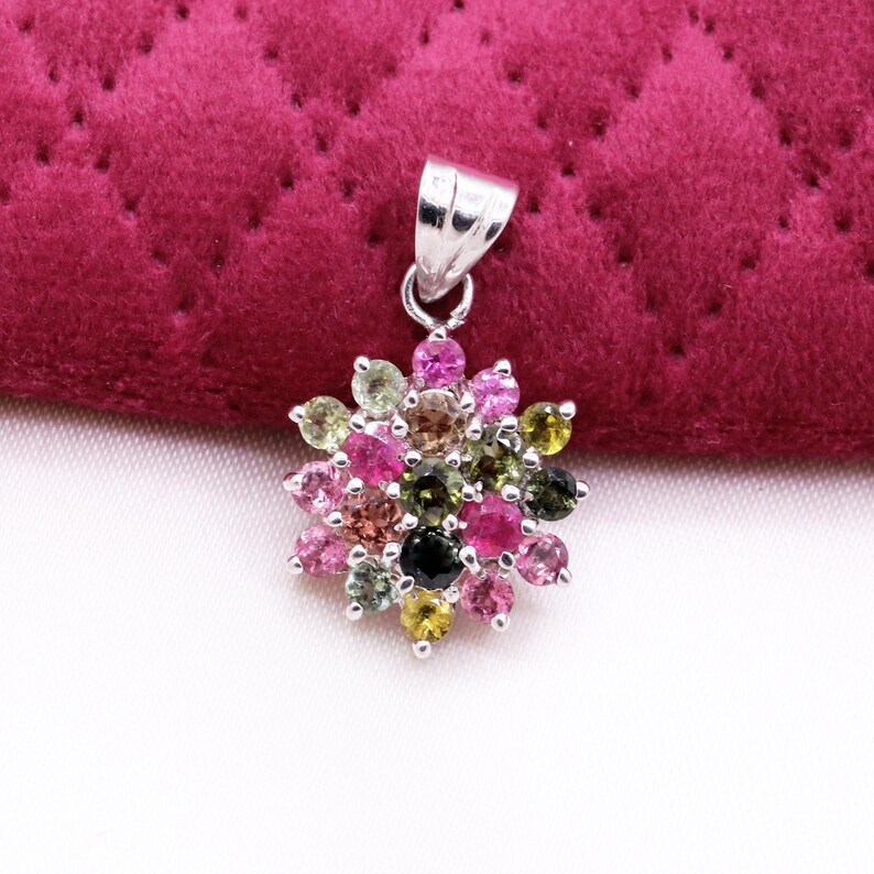 May include: A silver pendant with a cluster of colorful gemstones. The gemstones are arranged in a flower-like pattern, featuring shades of pink, green, yellow, and black. The pendant is set against a deep red, quilted background.