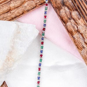 May include: A delicate tennis bracelet featuring a repeating pattern of red, green, and blue gemstones. The bracelet is made of silver and has a polished finish.