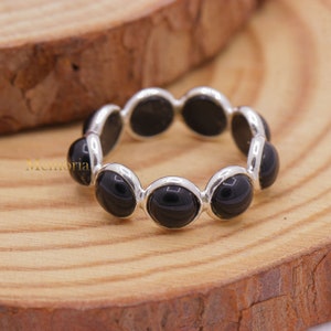 May include: A silver ring featuring a series of oval, glossy black stones. The stones are set in a continuous band, creating a sleek and modern design. The ring is displayed on a wooden surface.