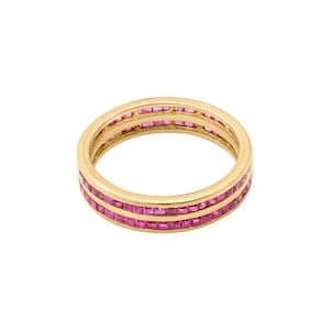 May include: A gold ring with three rows of square-cut pink gemstones. The ring is set against a white background, showcasing the bright gold and the vibrant pink stones. The ring's design is simple yet elegant.