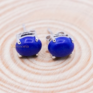 Könnte beinhalten: Ein Paar Ohrstecker mit ovalen, tiefblauen Edelsteinen, die in silberfarbenem Metall gefasst sind. Die Ohrringe werden auf einer hellen Holzoberfläche präsentiert. Die Ohrringe haben ein klassisches, elegantes Design.