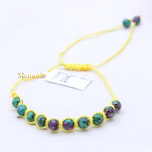Pode incluir: Um bracelete de pedras preciosas verdes e roxas com um cordão amarelo. O bracelete é ajustável com um nó deslizante.