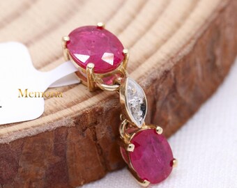 Puede incluir: Un colgante de oro con dos piedras preciosas de rubí de forma ovalada y un pequeño acento de diamante. El colgante está sobre una superficie de madera.