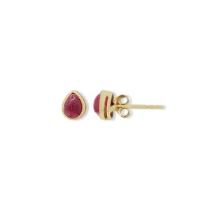 May include: A pair of stud earrings featuring teardrop-shaped, deep pink cabochon stones set in polished yellow gold bezels. One earring is shown from the front, while the other is viewed from the side, revealing the gold post and butterfly back.