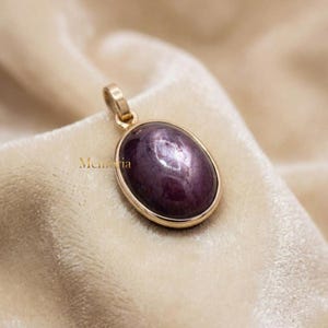 May include: An oval-shaped pendant featuring a deep purple star sapphire cabochon set in a polished gold-toned metal frame with a small bail at the top. The word Memoria is printed in small gold letters to the left of the pendant.
